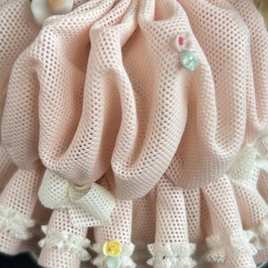 May include: Close-up of a porcelain figurine's dress. The dress is a light pink, open-weave fabric with a ruffled hem and a white bow. Small floral accents and a heart-shaped embellishment add detail to the design.