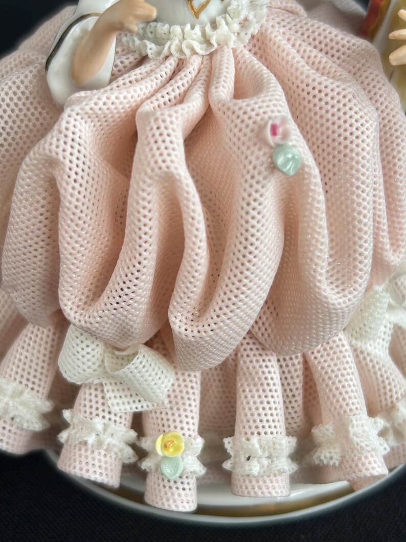 May include: Close-up of a porcelain figurine's dress. The dress is a light pink, open-weave fabric with a ruffled hem and a white bow. Small floral accents and a heart-shaped embellishment add detail to the design.
