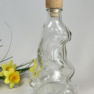 Könnte beinhalten: Eine klare Glasflasche in Hasenform mit einem Holzstopfen. Die Flasche hat ein detailliertes Kaninchen-Design. Die Flasche ist leer und steht auf einer weißen Oberfläche. Gelbe Blumen sind im Hintergrund.