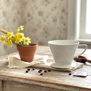 Könnte beinhalten: Ein weißer Melitta-Kaffeefilter aus Keramik mit dem Text "Melitta 102" liegt auf einem weißen Tuch. Ein kleiner Terrakotta-Topf enthält gelbe Blumen. Kaffeebohnen sind auf einem alten Holztisch verstreut.