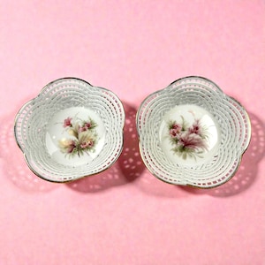 Könnte beinhalten: Zwei weiße Porzellanschalen mit floralem Design und einem filigranen, durchbrochenen Rand. Die Schalen haben einen goldenen Rand und sind auf einem rosa Hintergrund platziert. Das florale Design zeigt rosa und grüne Blumen.