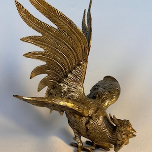 Könnte beinhalten: Eine detaillierte Bronzestatue eines Hahns mit ausgebreiteten Flügeln. Die Skulptur hat aufwendige Federdetails und eine strukturierte Oberfläche. Der Hahn ist in Aktion dargestellt, mit gesenktem Kopf und offenem Schnabel, was auf ein Krähen hindeutet.