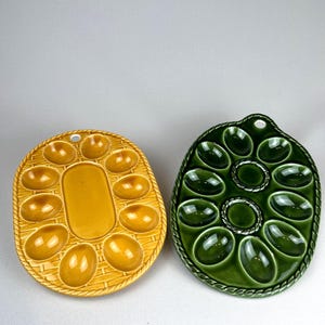May include: Two ceramic egg serving platters. One is oval-shaped, yellow, and has a basket weave design. The other is green, with a decorative rope-like border. Each platter has egg-shaped indentations.