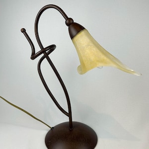 May include: A decorative table lamp with a bronze-colored metal base and stem. The lamp features a curved design with a flower-shaped shade in a pale yellow color. The lamp has a cord for power.