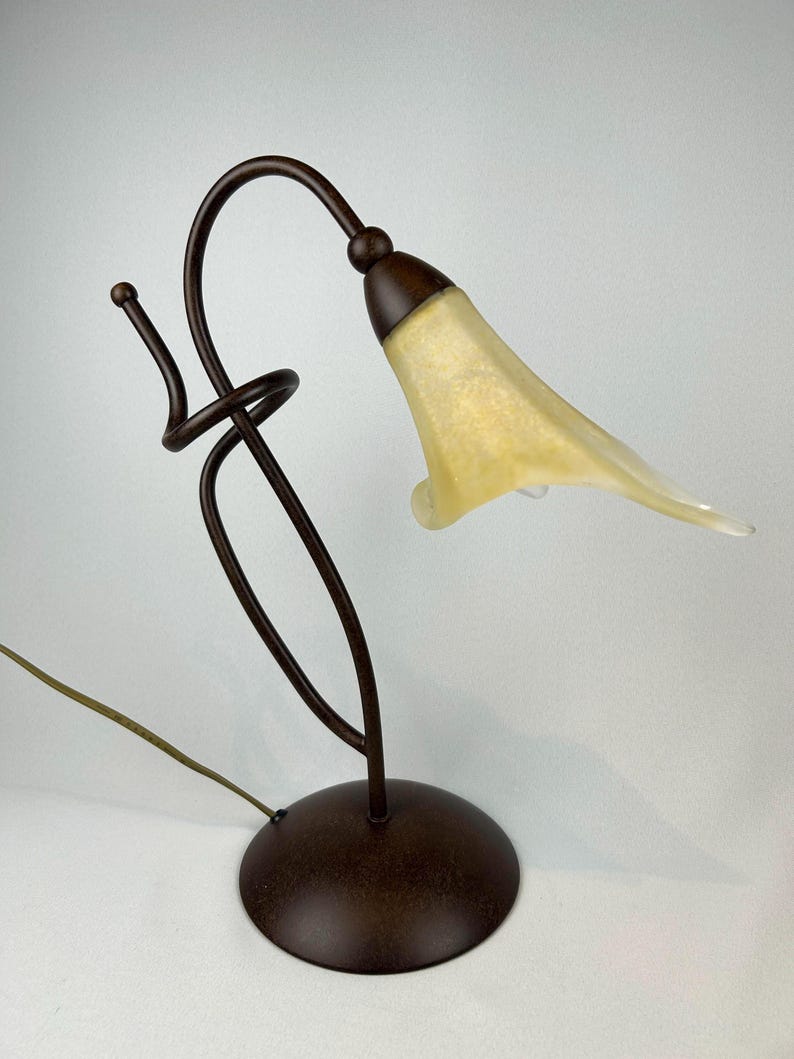May include: A decorative table lamp with a bronze-colored metal base and stem. The lamp features a curved design with a flower-shaped shade in a pale yellow color. The lamp has a cord for power.