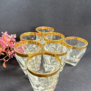 Könnte beinhalten: Set aus sieben klaren, facettierten Trinkgläsern mit Goldrand. Die Gläser sind auf einer dunklen Oberfläche angeordnet, mit einer kleinen Anordnung rosa Blumen im Hintergrund. Die Gläser haben ein klassisches, elegantes Design.