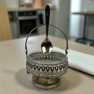 May include: A silver sugar bowl with a clear glass insert and a decorative filigree design. A silver spoon with a red handle rests in the bowl's handle. The bowl sits on a table.
