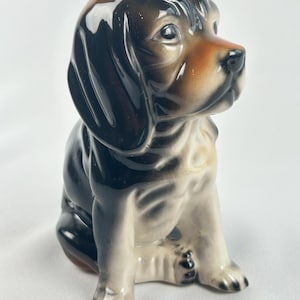 Peut inclure: Une figurine en céramique brillante représentant un chiot assis avec de longues oreilles tombantes. Le chien présente un pelage tricolore avec des marques noires, fauves et blanches, une finition émaillée lisse et un regard tourné vers la droite.