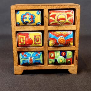 May include: A small wooden cabinet with six colorful drawers. Each drawer is rectangular and features a unique hand-painted design with bright colors like yellow, red, blue, and green. The cabinet is made of light brown wood.
