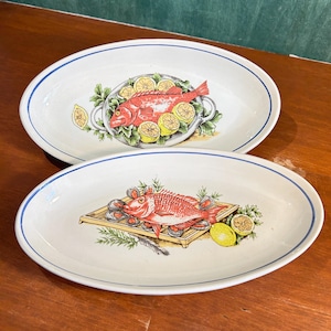 Könnte beinhalten: Zwei ovale Servierplatten mit weißem Grund und blauem Rand. Jede Platte zeigt eine lebendige Illustration eines roten Fisches, umgeben von Zitronen und Kräutern. Ein Fisch befindet sich auf einer silbernen Platte, der andere auf einem Holztablett.