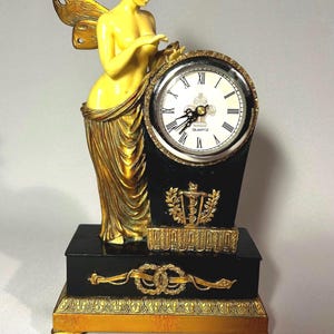 Könnte beinhalten: Eine dekorative Quarzuhr mit einer Statue einer Frau mit Flügeln. Die Uhr hat einen schwarzen Sockel mit goldenen Akzenten und ein weißes Zifferblatt mit römischen Ziffern. Die Statue ist hellgelb.