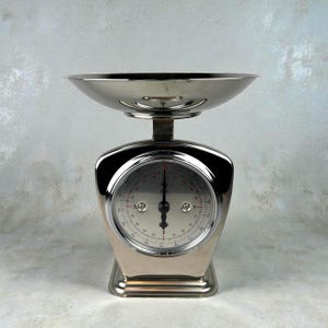 Op de afbeelding: Een chromen keukenweegschaal in vintage-stijl met een ronde wijzerplaat en een komvormige bovenkant. De wijzerplaat heeft zwarte en rode markeringen en de weegschaal heeft een stevige basis. Deze klassieke weegschaal is ontworpen voor het afwegen van ingrediënten.