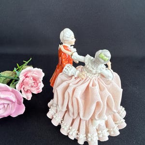 May include: A porcelain figurine depicts a couple in period attire dancing. The man wears a red coat, while the woman has a pink dress with white lace trim. Pink roses are in the foreground. The background is black.