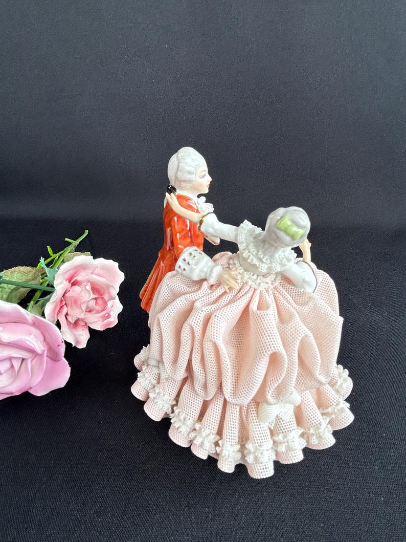 May include: A porcelain figurine depicts a couple in period attire dancing. The man wears a red coat, while the woman has a pink dress with white lace trim. Pink roses are in the foreground. The background is black.