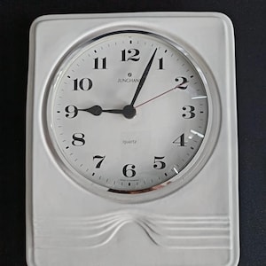 Könnte beinhalten: Eine rechteckige Wanduhr aus weißer Keramik mit abgerundeter Oberseite und einem dekorativen horizontalen Rillendetail am unteren Rand. Das Zifferblatt zeigt schwarze arabische Ziffern, schwarze Zeiger, einen roten Sekundenzeiger und JUNGHANS.