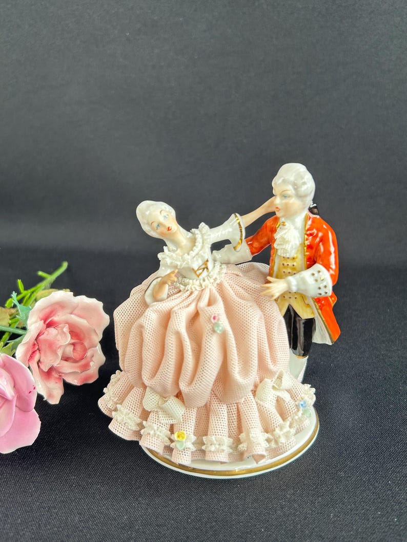 May include: A porcelain figurine depicting a couple in period attire dancing. The woman wears a pink dress with lace trim, while the man is in a red coat. The base is white with gold trim, and the figurine is set against a black background.