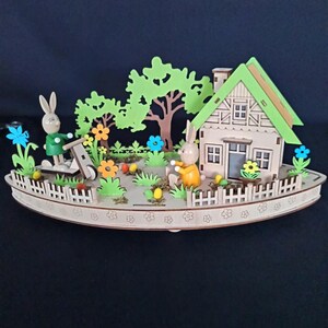 Könnte beinhalten: Oster-Diorama aus Holz mit einem Haus, Bäumen, Blumen und zwei Hasenfiguren. Das Haus hat ein grünes Dach und ein kleiner Zaun umgibt den Garten. Die Hasen tragen grüne und orangefarbene Outfits.