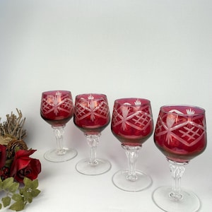 May include: Four ruby red wine glasses with etched floral and geometric designs. The glasses have clear stems and bases. The background is white, and the glasses are arranged in a row. A decorative arrangement of roses is in the background.