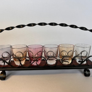 May include: A black metal caddy with a twisted handle and scrollwork base holding six small glass tumblers. The glasses feature varied translucent tints including clear, light pink, and amber tones, arranged in a single row within the frame.