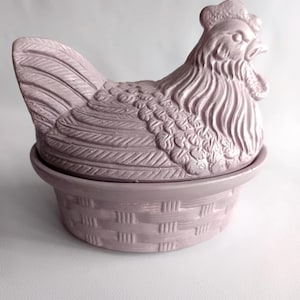 Puede incluir: Un recipiente de cerámica con tapa en color lavanda claro con forma de gallina sobre un nido. La tapa presenta una gallina moldeada detallada con plumas texturizadas, mientras que la base está diseñada como una cesta tejida con acabado mate.
