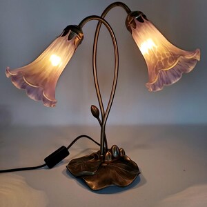 May include: A decorative table lamp featuring two flower-shaped shades in shades of lavender and white. The lamp has a bronze-colored metal base with a leaf design and curved stems. The lamp is plugged into a wall outlet.