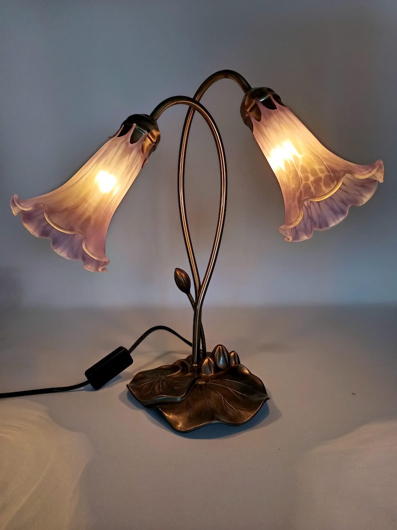 May include: A decorative table lamp featuring two flower-shaped shades in shades of lavender and white. The lamp has a bronze-colored metal base with a leaf design and curved stems. The lamp is plugged into a wall outlet.
