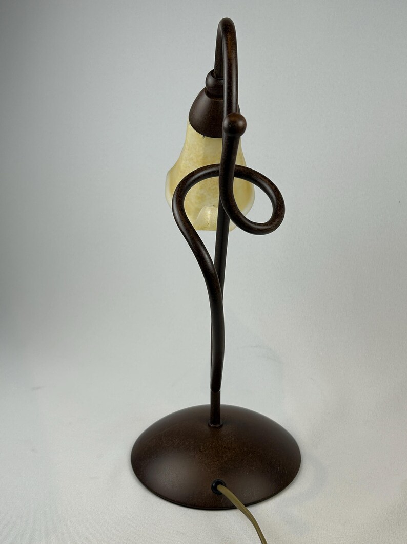 May include: A decorative table lamp with a bronze finish. The lamp features a curved stem and a swirling design element. The lampshade is a teardrop shape with a cream-colored glass. The lamp has a round base and a power cord.