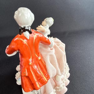 May include: A porcelain figurine depicting a couple dancing. The man wears a bright orange coat with gold accents and a black bow tie. The woman is in a pink dress with lace trim. The base is white with a gold rim.