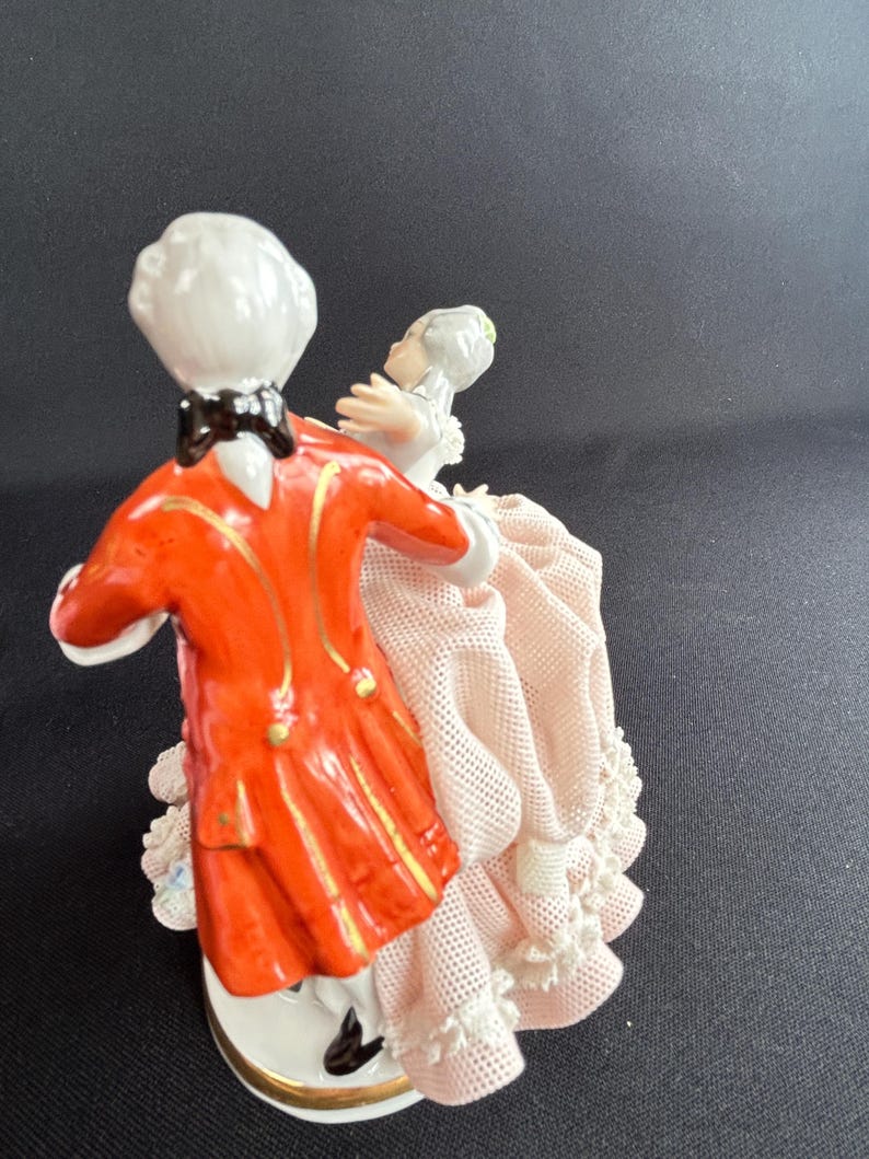 May include: A porcelain figurine depicting a couple dancing. The man wears a bright orange coat with gold accents and a black bow tie. The woman is in a pink dress with lace trim. The base is white with a gold rim.