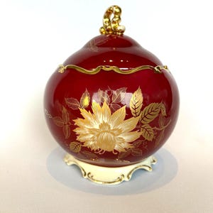 Könnte beinhalten: Eine rote Keramikdose mit Deckel, verziert mit goldenen Blumenmustern und Verzierungen. Die Dose steht auf einem cremefarbenen Sockel mit goldenen Details. Der Deckel hat einen goldenen Griff. Höhe ca. 15 cm.