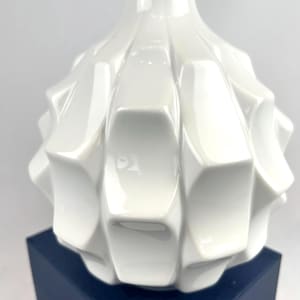 May include: A white ceramic vase with a geometric, textured design. The vase has a wide, rounded body that tapers to a narrow neck. The surface is glossy and reflects light. The vase is sitting on a dark blue box.