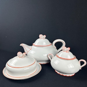 Könnte beinhalten: Ein weißes Keramik-Teeservice mit roter Verzierung, bestehend aus einer Teekanne, einer Zuckerdose und einer Butterdose mit Deckel. Jedes Teil hat ein rotes und weißes Blumendesign auf dem Deckel. Das Set wird vor dunklem Hintergrund präsentiert.