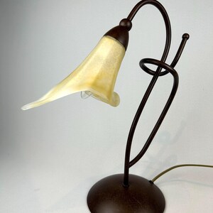 May include: A decorative table lamp with a unique design. The lamp features a brown metal base and stem, supporting a flower-shaped lampshade with a yellow and white color scheme. The lamp's design includes a curved stem and a spiral detail.