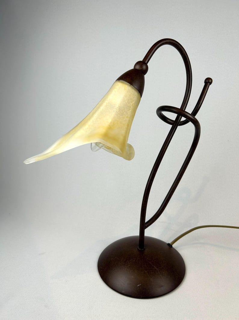 May include: A decorative table lamp with a unique design. The lamp features a brown metal base and stem, supporting a flower-shaped lampshade with a yellow and white color scheme. The lamp's design includes a curved stem and a spiral detail.