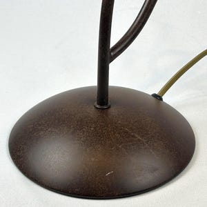 May include: A close-up of a brown metal lamp base with a circular design. The base has a textured finish and a dark brown color. A brown stem and a green cord are also visible.
