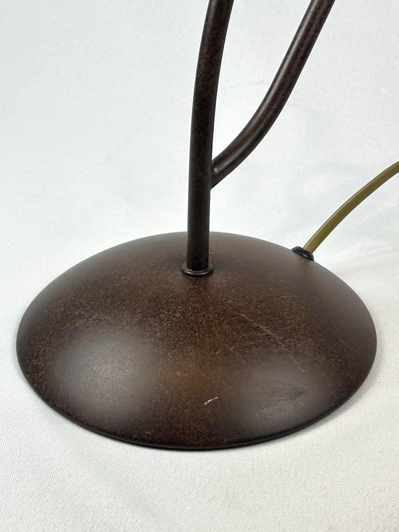 May include: A close-up of a brown metal lamp base with a circular design. The base has a textured finish and a dark brown color. A brown stem and a green cord are also visible.