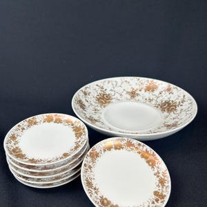 Könnte beinhalten: Eine Sammlung weißer Porzellangeschirrteile mit goldenem Blumenmuster. Das Set umfasst eine große Untertasse und mehrere kleinere Teller, die auf einer schwarzen Oberfläche gestapelt und angeordnet sind. Das goldene Design ist detailliert und bedeckt die Ränder der Teller.