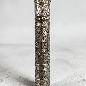 May include: A cylindrical silver-toned metal case featuring an intricate embossed pattern of swirling floral motifs and geometric Greek key borders. The object has a rounded, dome-shaped top and a matching rounded base, with a metallic finish.