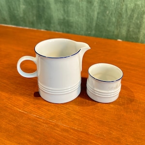 Könnte beinhalten: Weißer Keramik-Sahneaufgießer und kleine Tasse mit blauem Rand. Der Sahneaufgießer hat einen Griff und einen Ausguss. Beide Teile haben horizontale Rillen am Boden. Das Set steht auf einer Holzoberfläche.