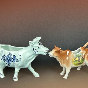 Peut inclure: Deux crémiers en forme de vache en céramique. L'un est bleu clair avec des motifs floraux bleus. L'autre est marron et blanc avec une scène de paysage peinte. Les deux ont une anse et un bec verseur.