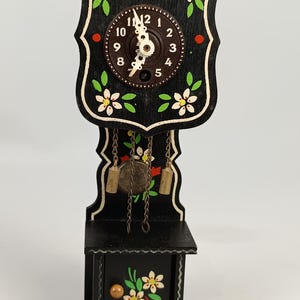 Puede incluir: Un pequeño reloj de madera negro con un diseño floral decorativo. La esfera del reloj es marrón con números y manecillas blancas. El reloj tiene un péndulo y dos pesas colgando de cadenas. El reloj está decorado con flores blancas y hojas verdes.