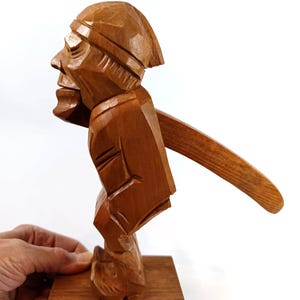 Könnte beinhalten: Ein hölzerner Nussknacker in Form eines Mannes. Der Nussknacker ist braun und steht auf einem rechteckigen Holzsockel. Der Hebel erstreckt sich von der Rückseite der Figur. Die Figur wird von einer Hand gehalten.