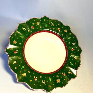 Könnte beinhalten: Ein dekorativer Weihnachtsteller mit gewelltem Rand. Der grüne Rand ist mit festlichen Illustrationen von Rentieren, Weihnachtsbäumen und Ornamenten verziert. Ein rotes Band umrandet die cremefarbene Mitte.