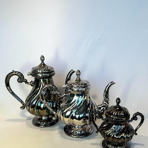 Könnte beinhalten: Drei antike silberne Teeservice-Teile. Das Set umfasst eine Teekanne, einen Sahnekännchen und eine Zuckerdose. Jedes Stück hat ein geriffeltes Design und verzierte Griffe. Die Teekanne hat einen gebogenen Ausguss und einen gewölbten Deckel mit einem Knauf.