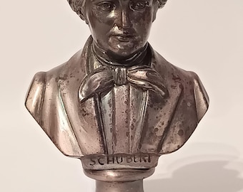 May include: A metallic silver-toned bust of a person with wavy hair, wearing a collared shirt and a bow tie. The base of the sculpture features the name SCHUBERT embossed in capital letters. The surface shows signs of aged patina and wear.