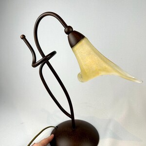 May include: A decorative table lamp with a bronze-colored metal base and stem. The lamp features a curved design with a yellow and white glass shade shaped like a flower. The lamp has a round base and a cord.