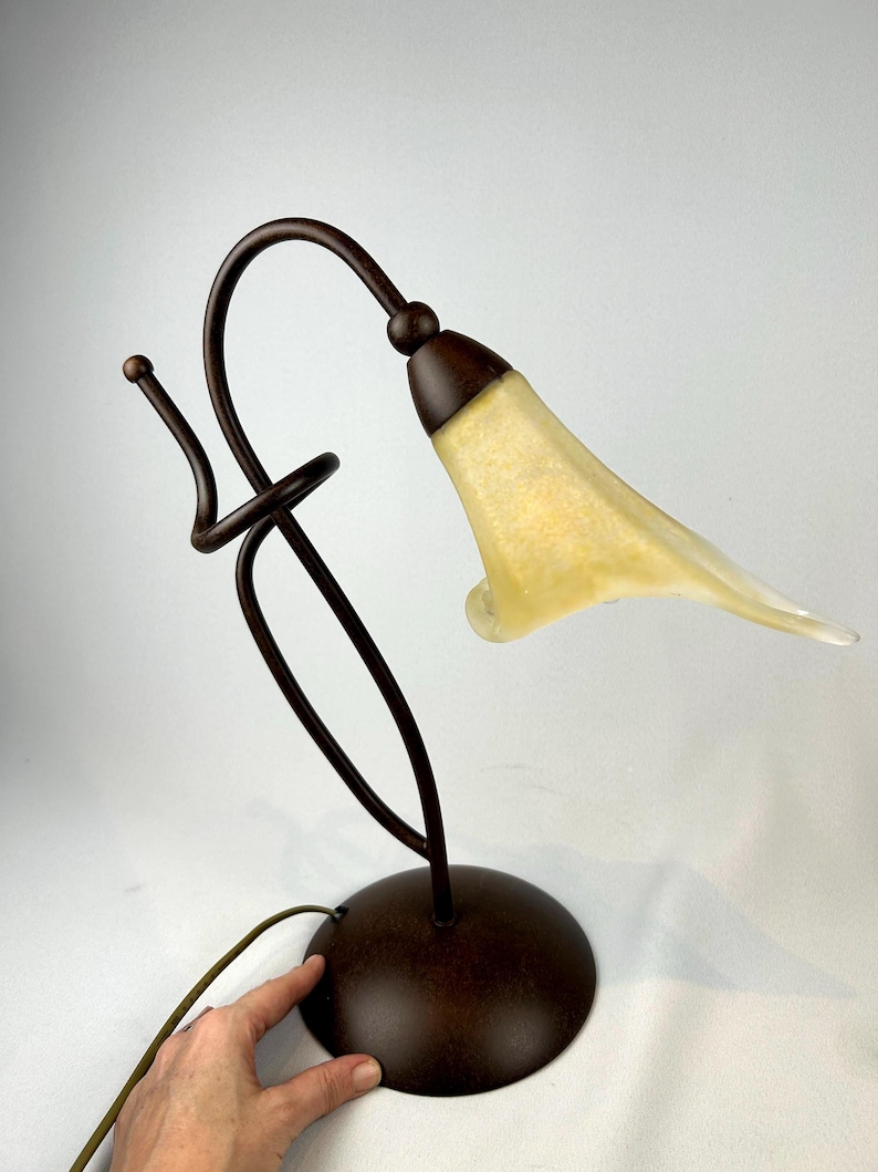 May include: A decorative table lamp with a bronze-colored metal base and stem. The lamp features a curved design with a yellow and white glass shade shaped like a flower. The lamp has a round base and a cord.