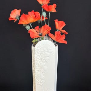 May include: A white, rectangular vase with embossed floral and face designs holds a bouquet of vibrant orange poppies. The poppies have green stems and buds, set against a black background.