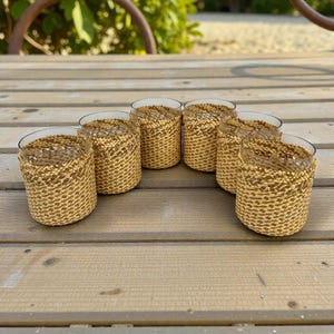 Puede incluir: Seis vasos transparentes con fundas tejidas de color beige. Los vasos están dispuestos en semicírculo sobre una superficie de madera. Las fundas tejidas añaden un elemento rústico y texturizado a la cristalería.