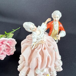 May include: A porcelain figurine set depicting a couple in 18th-century attire. The woman wears a pink dress with lace trim, while the man is in a red coat. The figures have detailed facial features and are posed as if dancing.